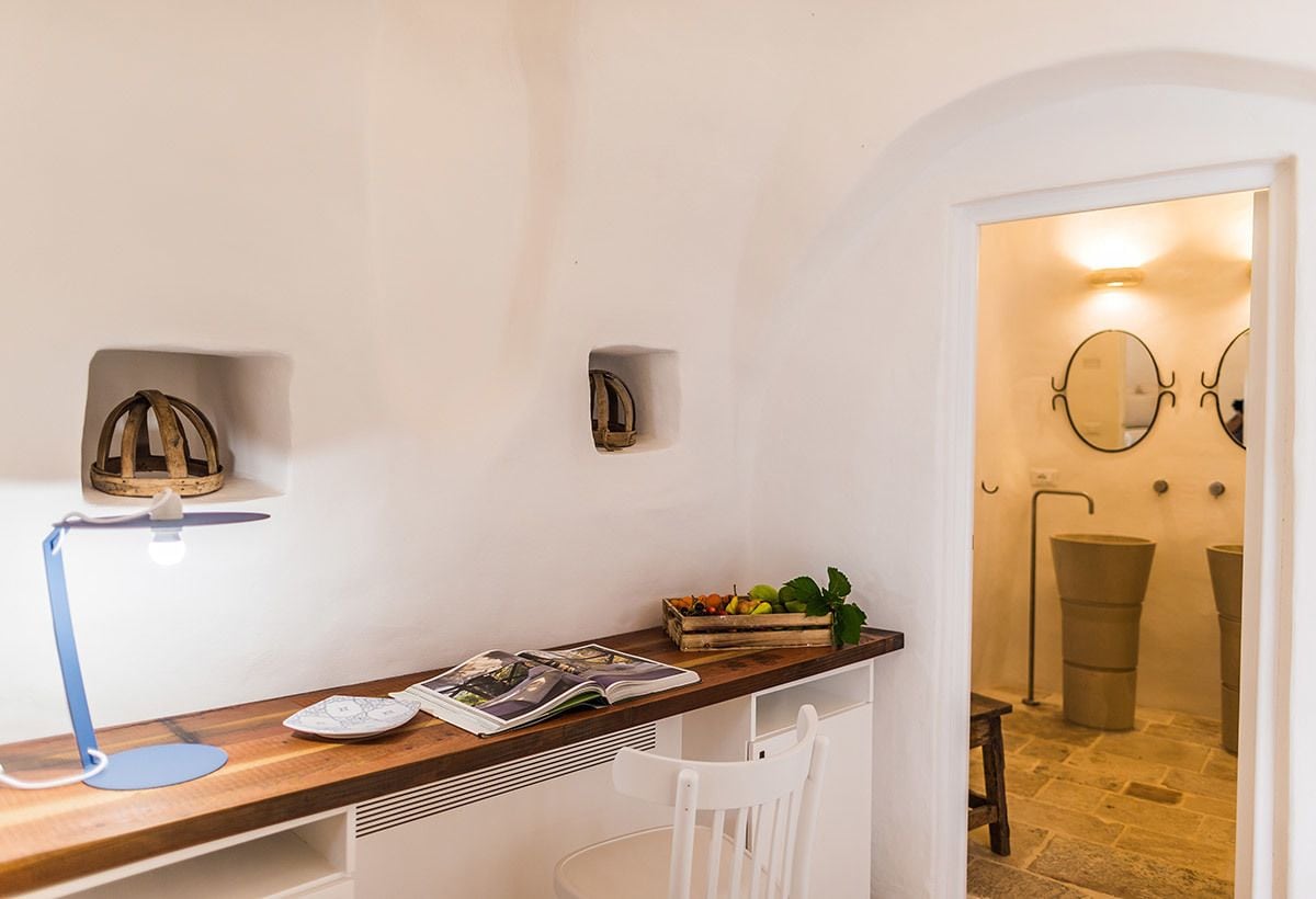 Una habitación pequeña y acogedora con un escritorio de madera, una silla blanca, una lámpara y un estante con objetos decorativos. Hay una puerta que da a un baño con espejo y un cubo de basura.