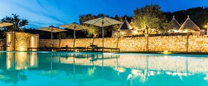 A swimming pool with stone walls and umbrellas, surrounded by stone buildings with conical roofs. - Borgo Canonica (Cisternino)