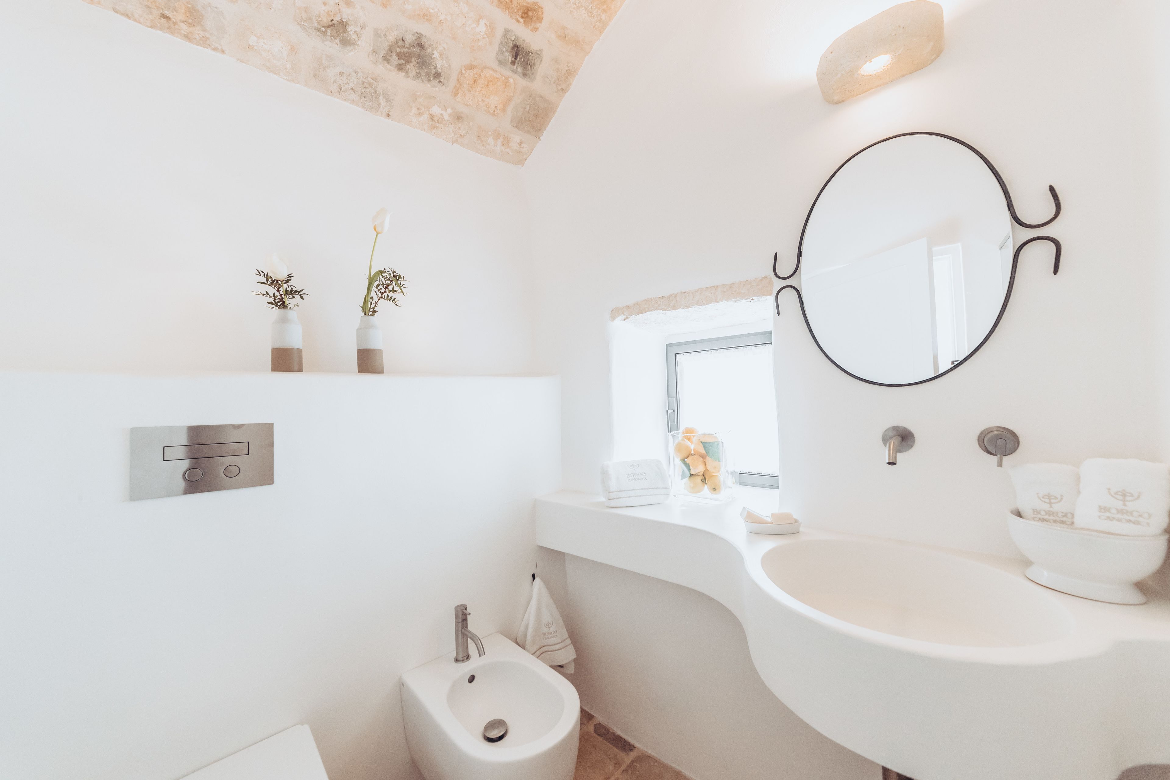 Casa de banho moderna com lavatório branco, um bidé e um espelho. Existem dois pequenos vasos com flores secas na parede, e uma parede de pedra acima do lavatório.