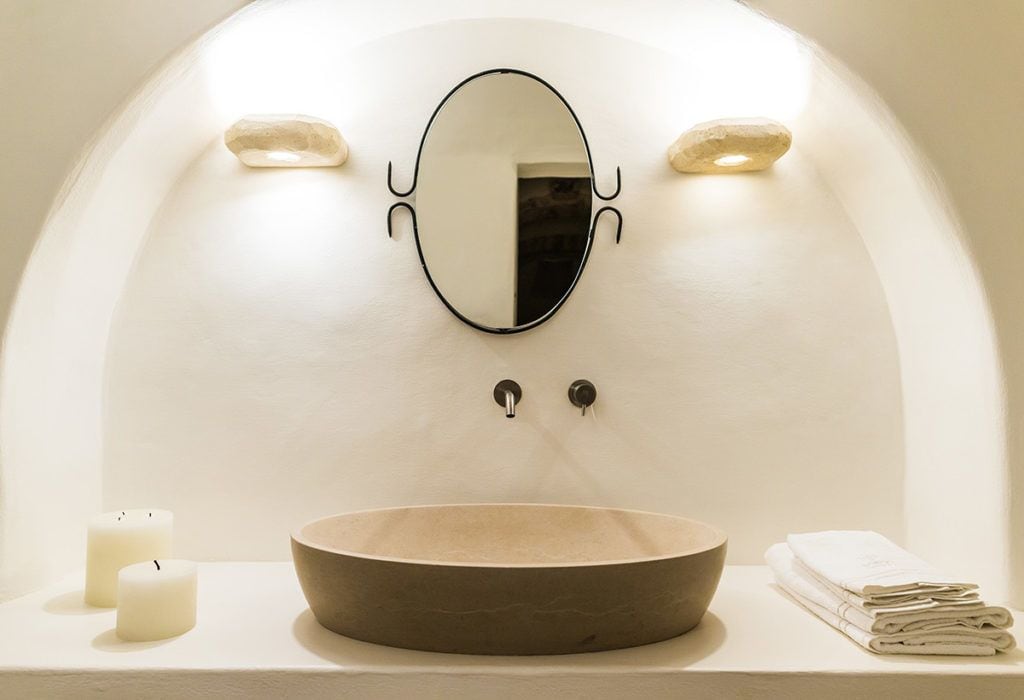 A bathroom with a round sink, an oval mirror, two wall-mounted lights, and two candles on the left.