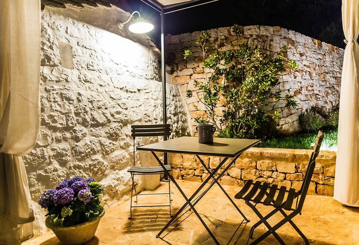 An outdoor patio with a stone wall, a table, two chairs, a potted plant, and a lamp.