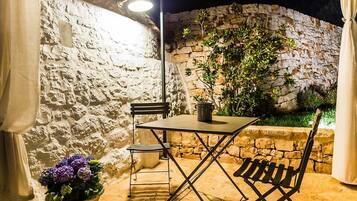 An outdoor patio with a stone wall, a table, two chairs, a potted plant, and a lamp.