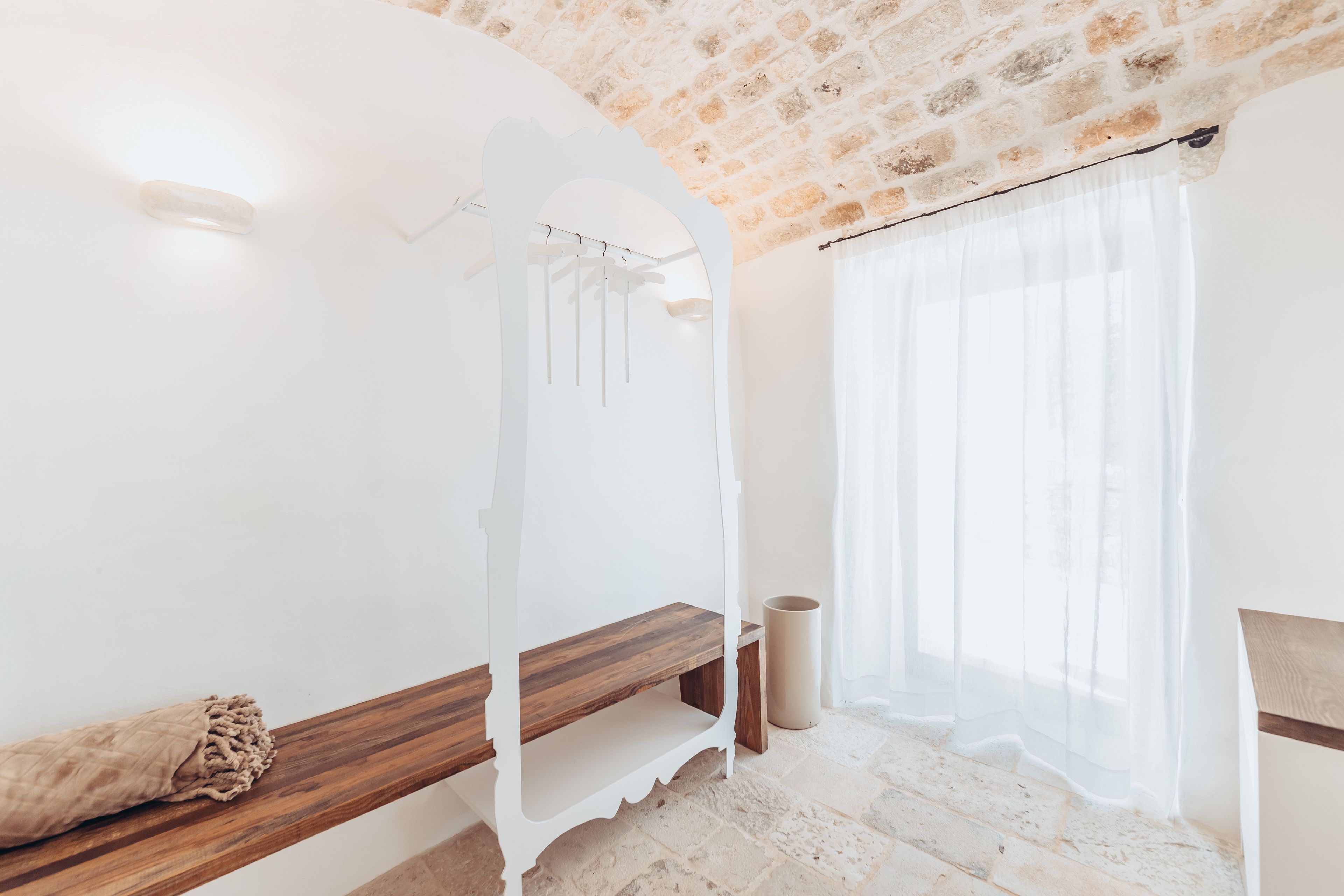 Una stanza con soffitto in pietra, pareti bianche, una panca di legno, un lampadario e una finestra con tende leggere.