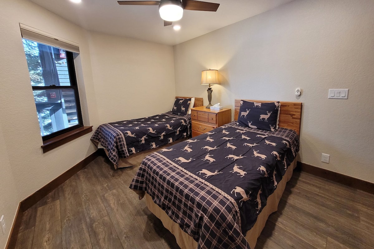 A bedroom with two beds, a ceiling fan, a window, a nightstand with a lamp, and a wooden floor.
