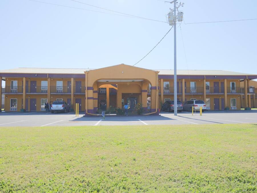 Un motel con entrada central, varios espacios de estacionamiento y un área verde al frente.