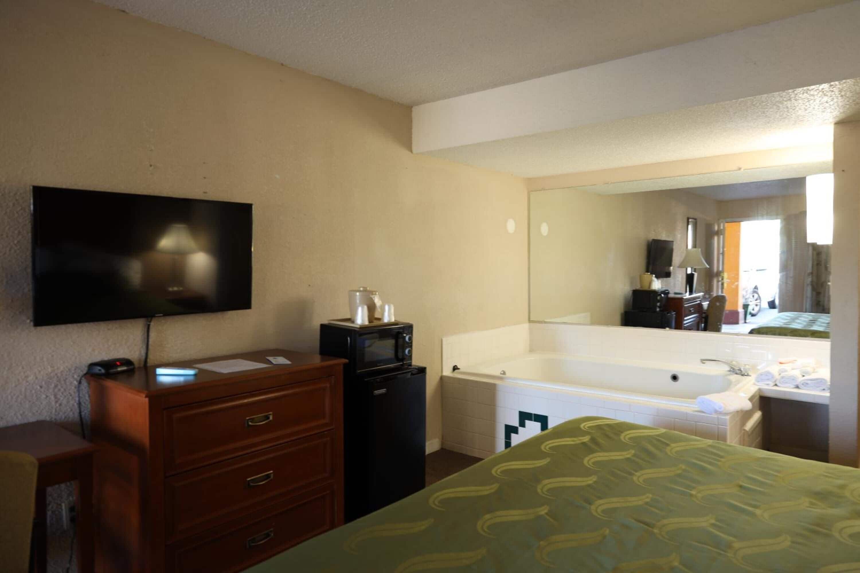 A hotel room with a flat-screen TV, a wooden dresser, a black refrigerator, a large bathtub, and a green patterned bedspread.