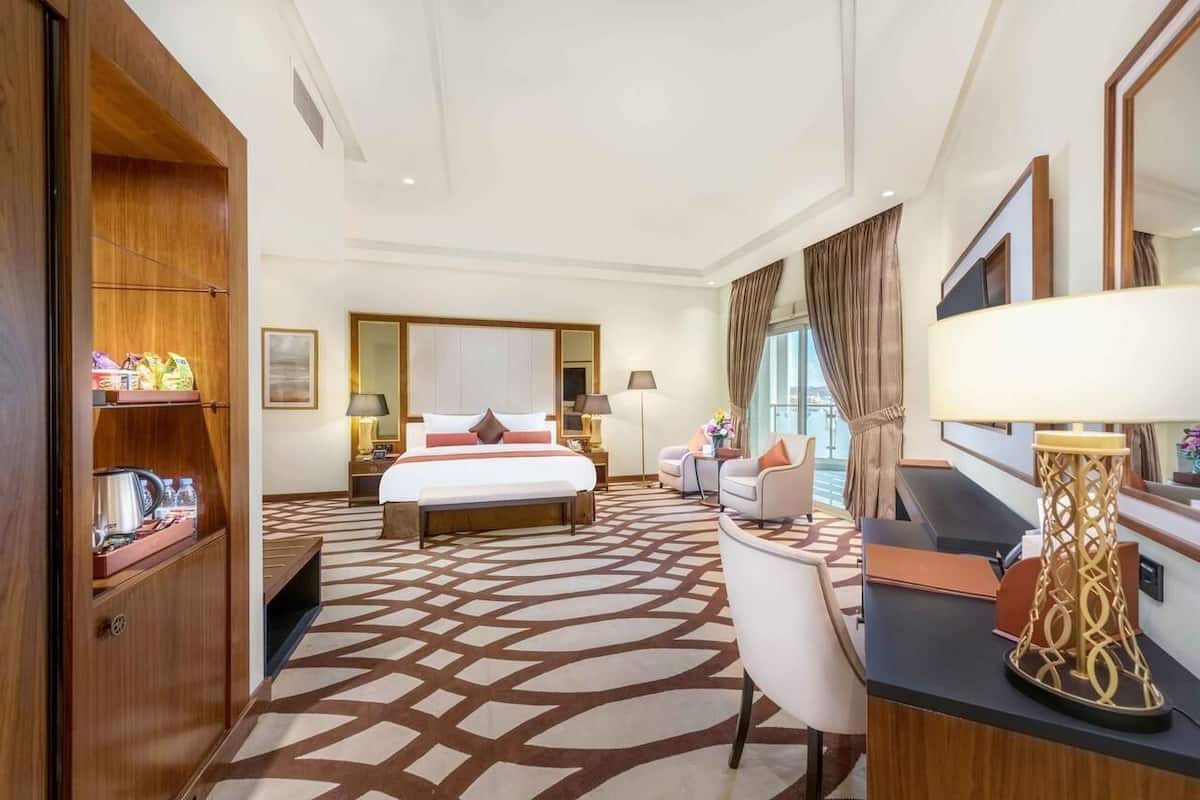 Une chambre d’hôtel avec un grand lit, un bureau avec une lampe, une chaise, une armoire et une grande fenêtre avec des rideaux.
