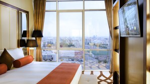 A hotel room with a large bed, a city view through floor-to-ceiling windows, a flat-screen TV mounted on the wall, and a modern lamp.
