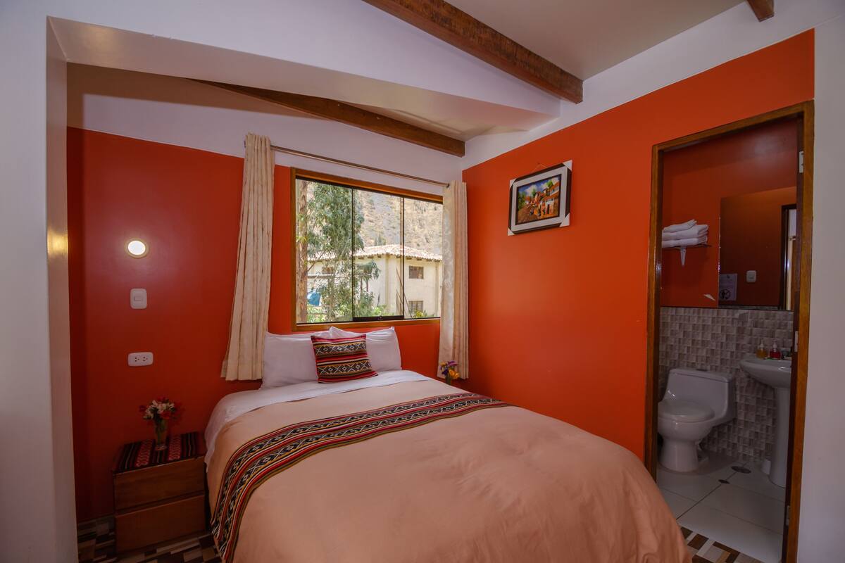 Une chambre à coucher avec un lit, une fenêtre avec des rideaux, une table de chevet avec un vase, et une salle de bain visible à travers une porte ouverte.