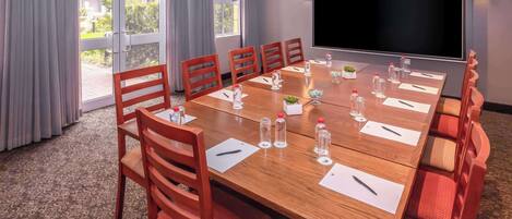 Uma sala de conferências com uma mesa grande, cadeiras, uma tela de projetor e vista para o exterior pelas janelas.