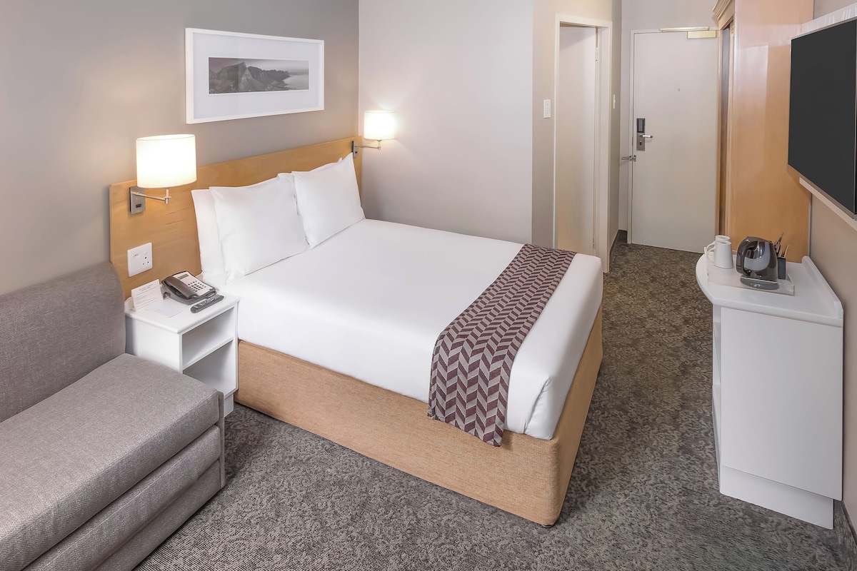 A hotel room with a bed, bedside tables, a desk, a chair, a TV, and a door.