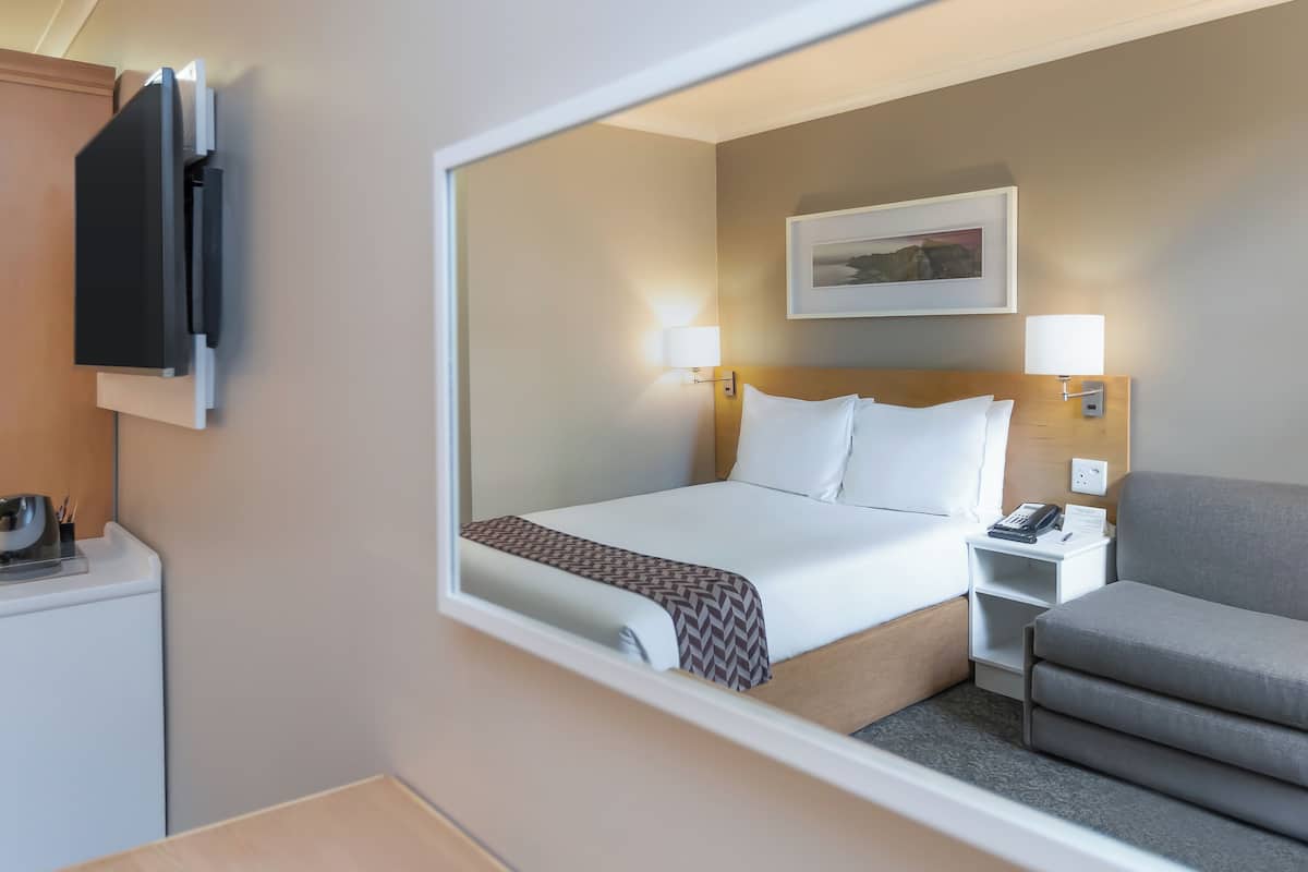 Habitación de hotel con cama, mesita de noche con teléfono, una silla, escritorio con lámpara y televisor en la pared.
