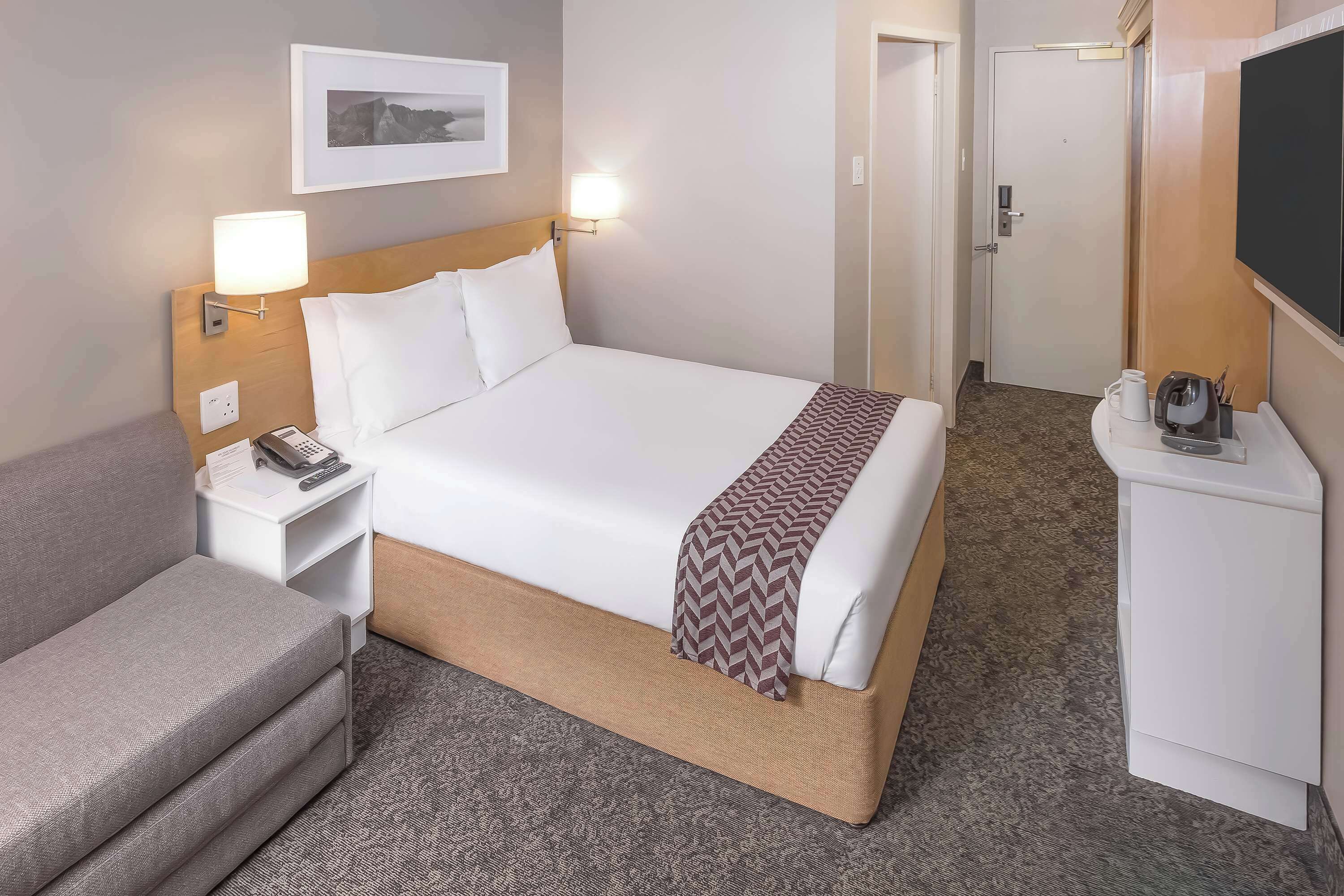 Une chambre d’hôtel comprenant un lit, une table de chevet avec une lampe, un petit bureau avec un téléphone, une télévision et un sol recouvert d’une moquette.