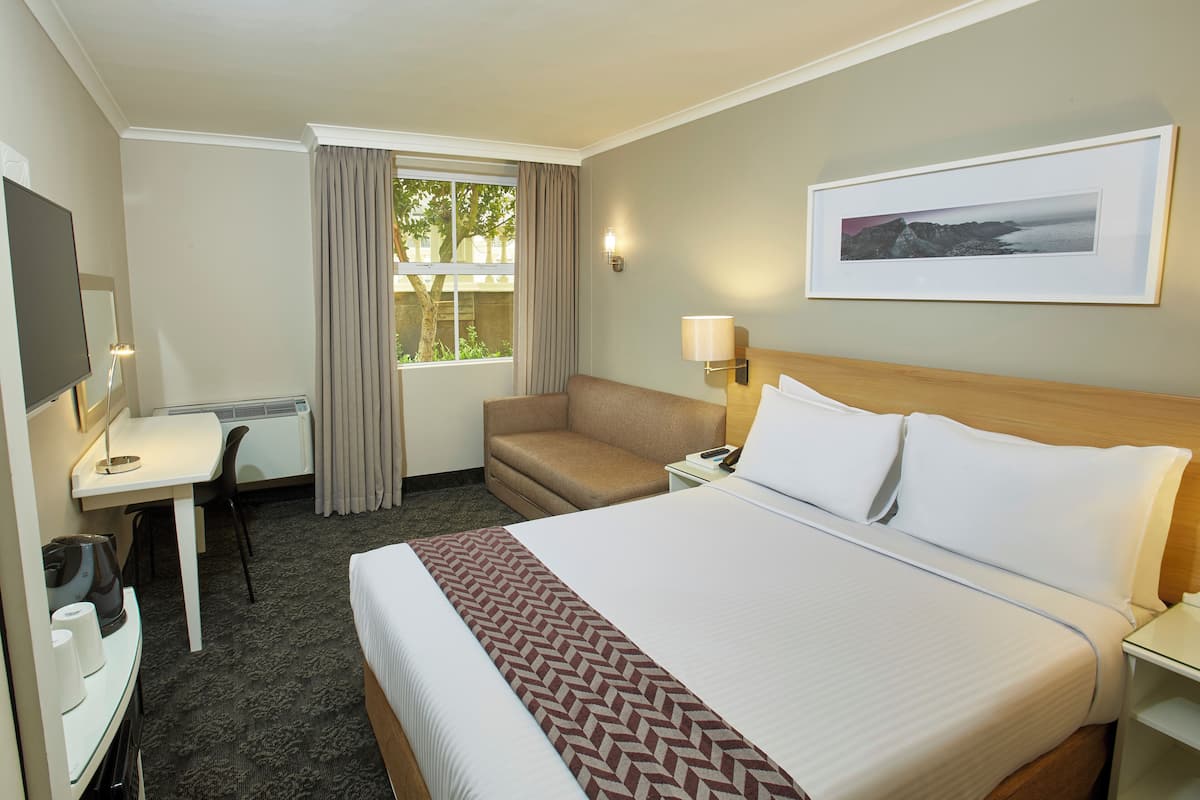 Habitación de hotel con cama, escritorio, silla, televisor, ventana con cortinas e imagen enmarcada en la pared.