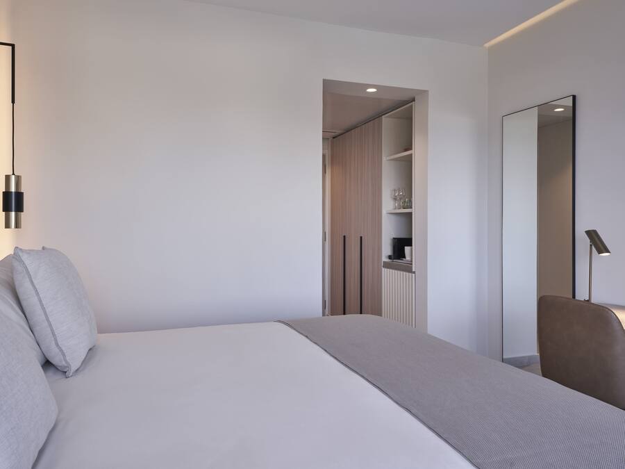 A modern hotel room with a large bed, a bedside table, a chair, a wardrobe, and a mirror.
