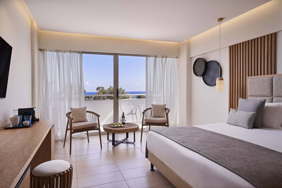 Een hotelkamer met een bed, twee stoelen, een tafeltje, een bureau en uitzicht op zee.