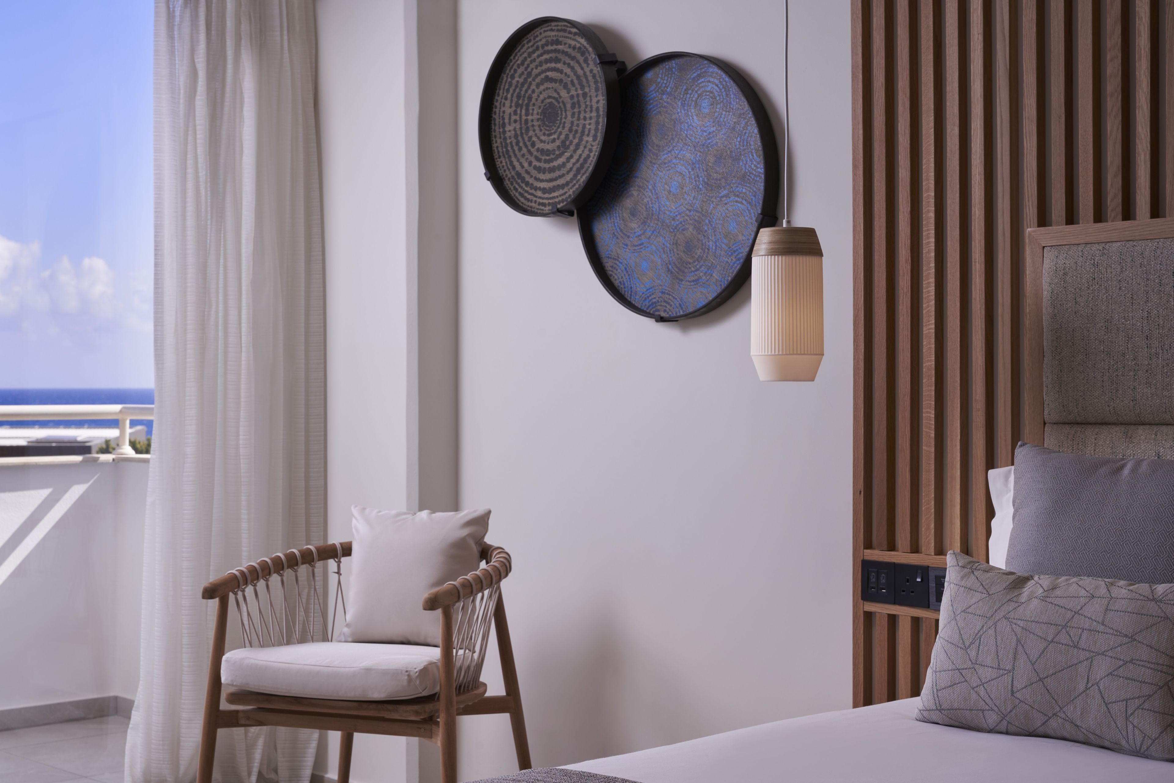 A hotel room with a bed, a chair, a wall-mounted lamp, and a view of the sea.