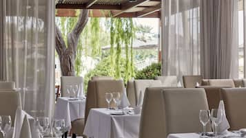 Restaurante com mesas preparadas para refeições, oferecendo vista para áreas verdes e uma árvore lá fora.