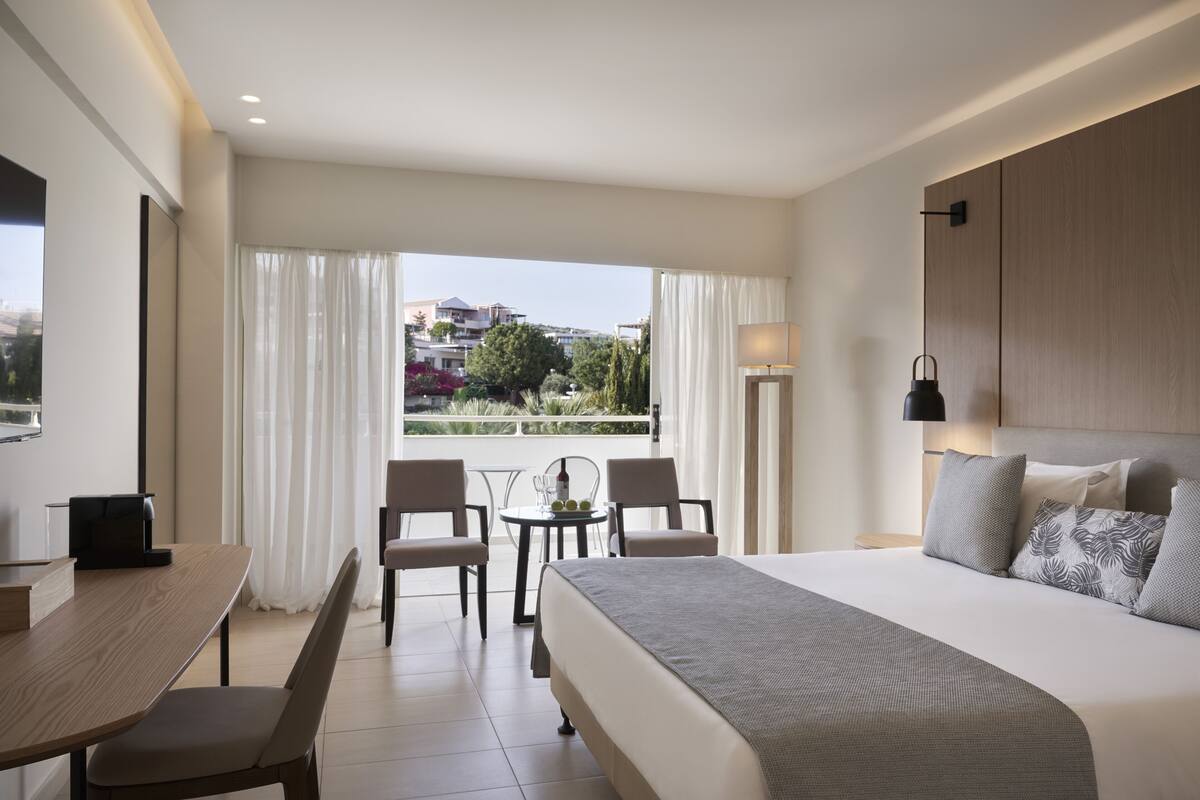 Een moderne hotelkamer met een groot bed, een bureau met een stoel, een kleine tafel met twee stoelen, en een balkon met uitzicht op de stad.