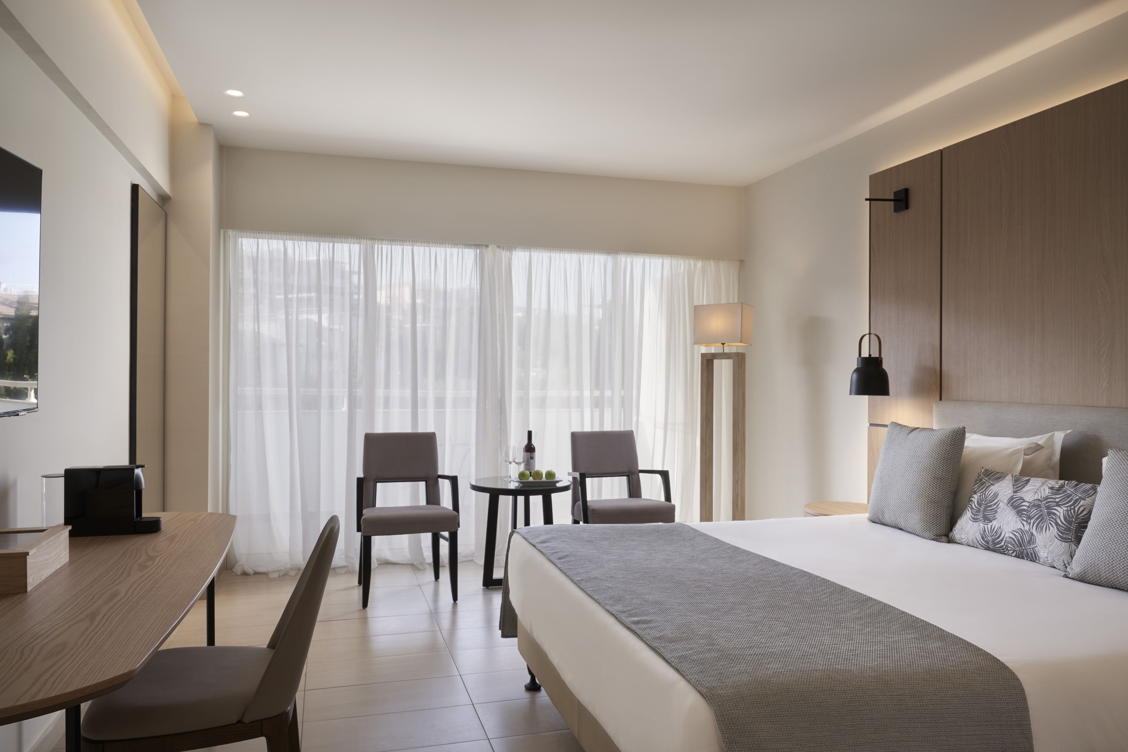 Une chambre d’hôtel avec un lit, deux chaises, une petite table, une lampe et une vue sur la ville à travers de fines rideaux.