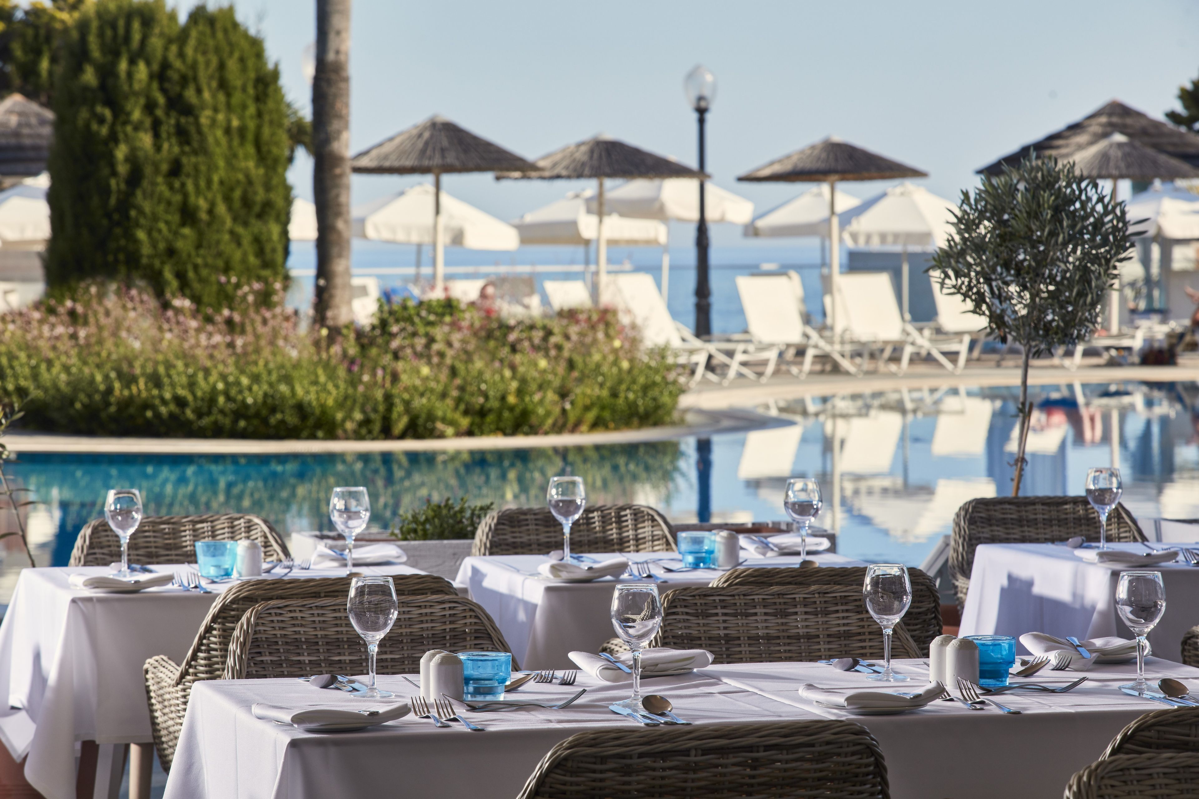 A poolside dining area with tables set for two, overlooking a pool with lounge chairs and umbrellas.