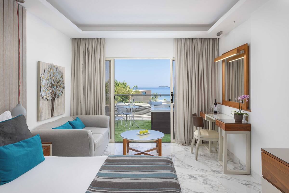 Een hotelkamer met een bed, een bank, een eettafel, een stoel, een bureau en een groot raam met uitzicht op zee en een gebouw.