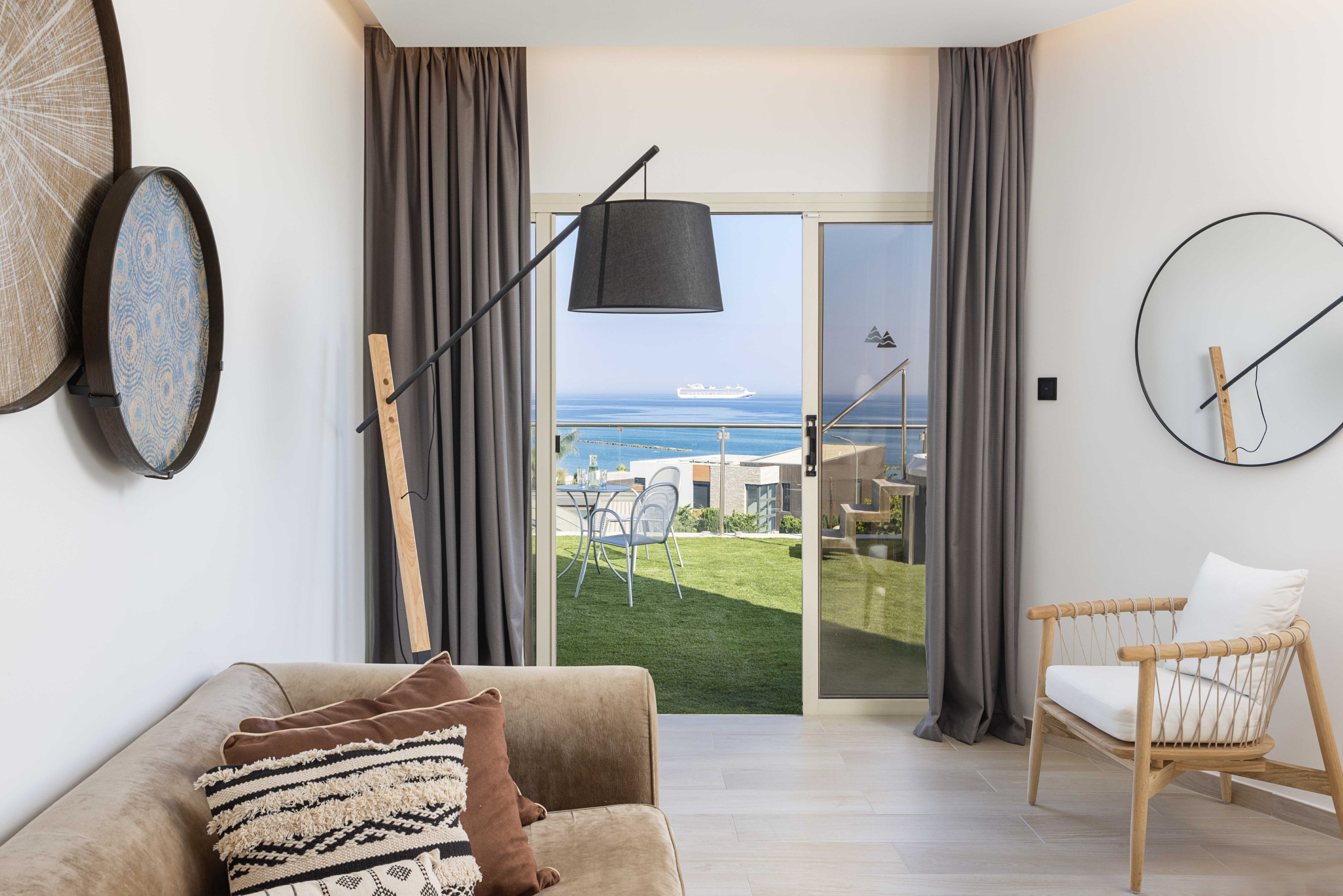 A modern living room with a sofa, a chair, a large window with curtains, a wall clock, and a view of the ocean.