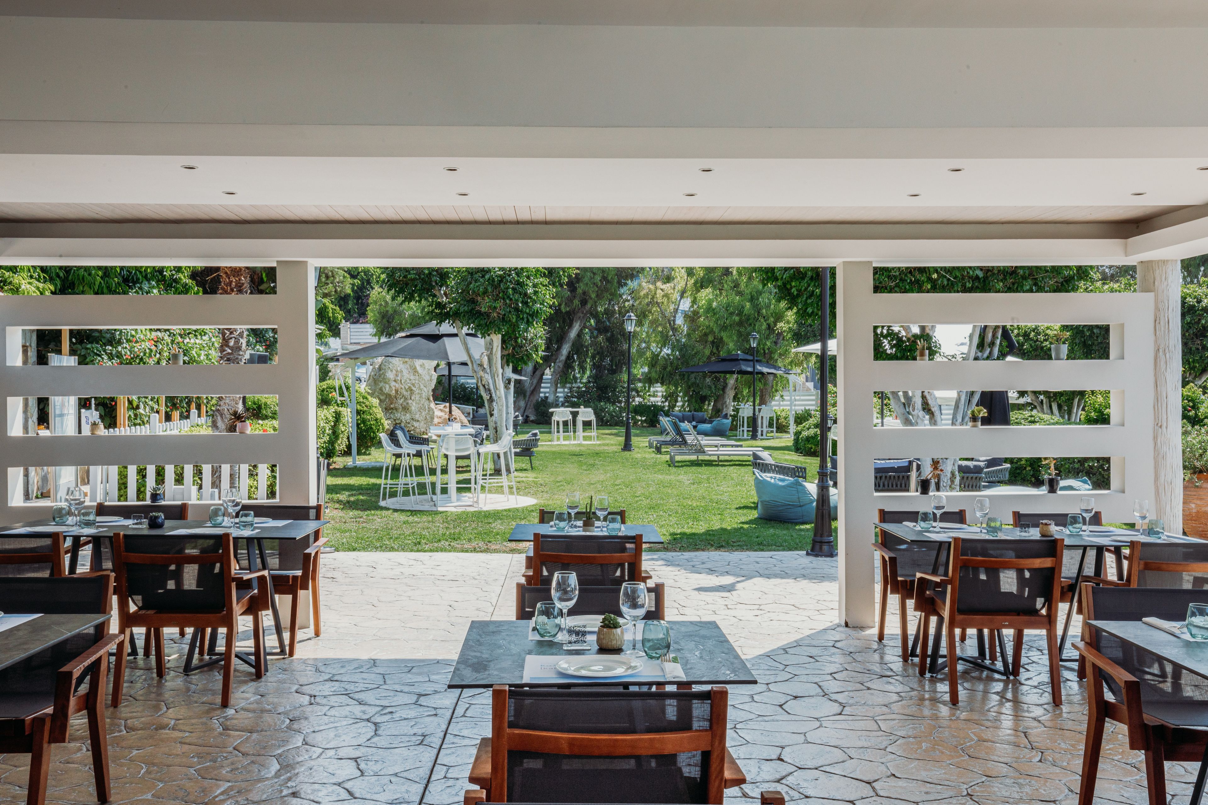 Un área de comedor al aire libre con mesas y sillas, vista a un jardín con sombrillas, y una estructura de valla blanca.