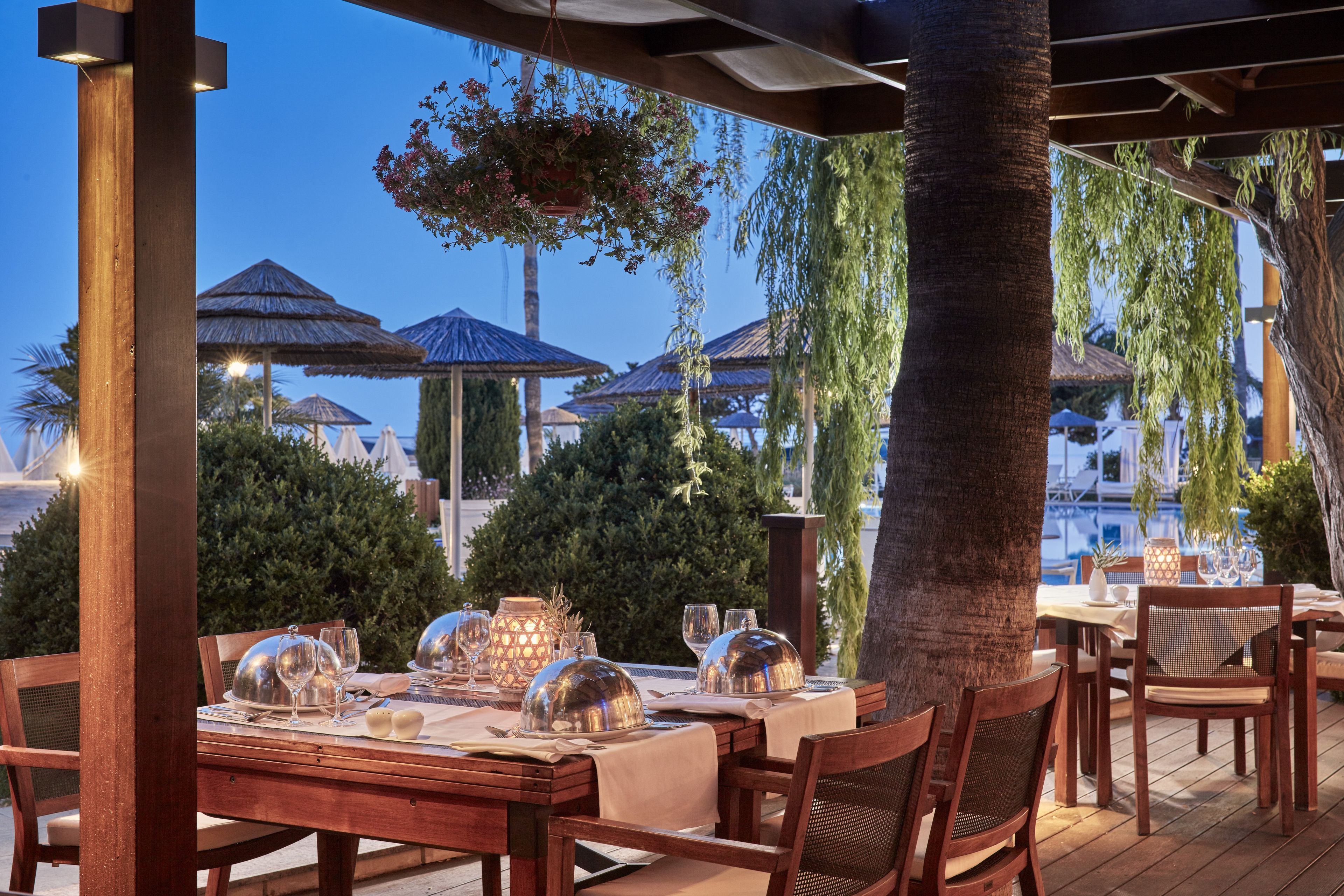 An outdoor dining area with tables set for a meal, surrounded by wooden pillars and covered with a pergola.