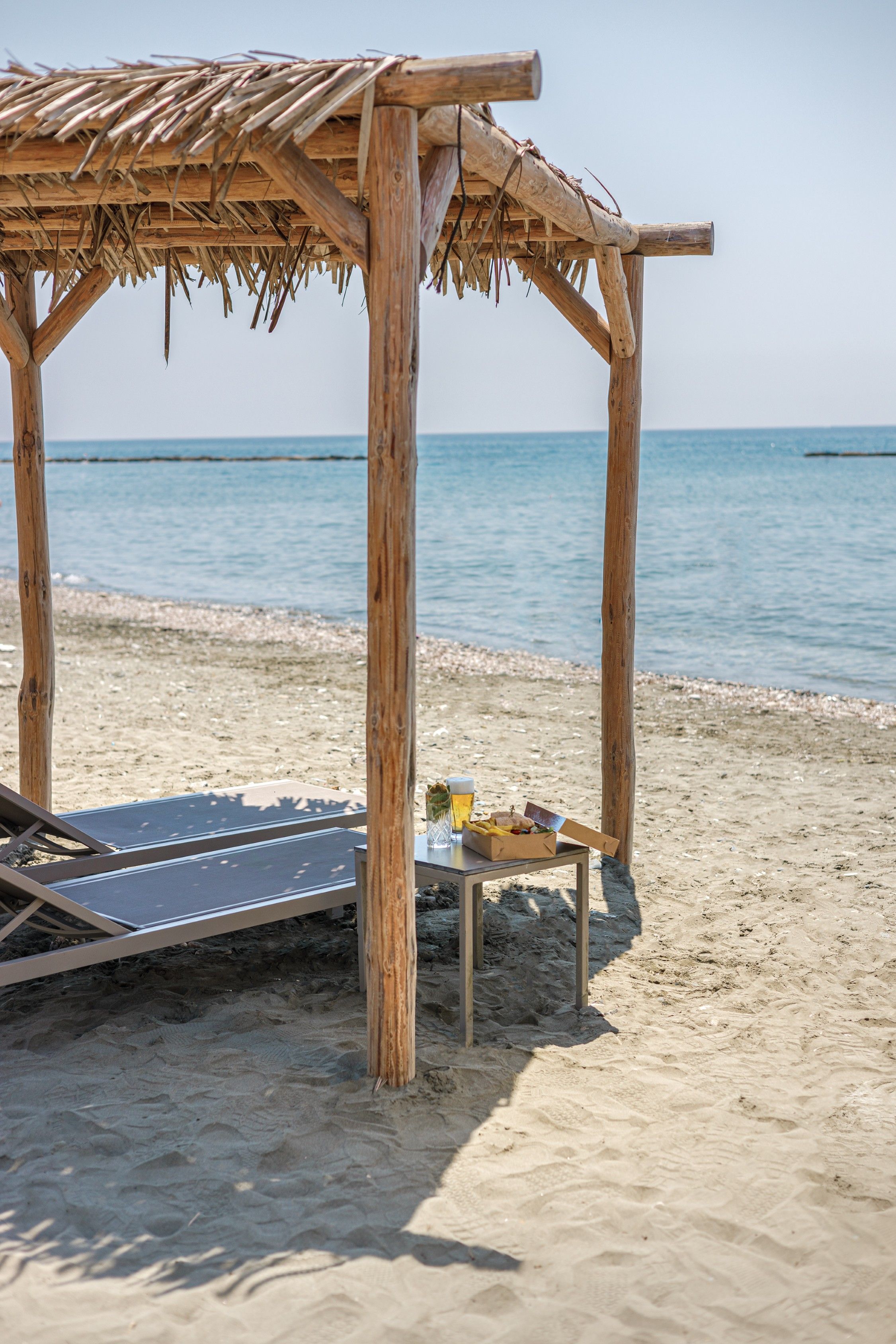 A wooden beach hut with a thatched roof, a lounge chair, and a table with a drink and sandwich.