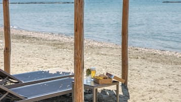 Una cabaña de playa de madera con techo de paja, una silla de descanso y una mesa con una bebida y un sándwich.