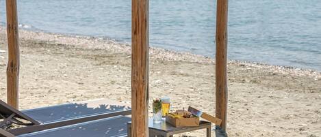 A wooden beach hut with a thatched roof, a lounge chair, and a table with a drink and sandwich.