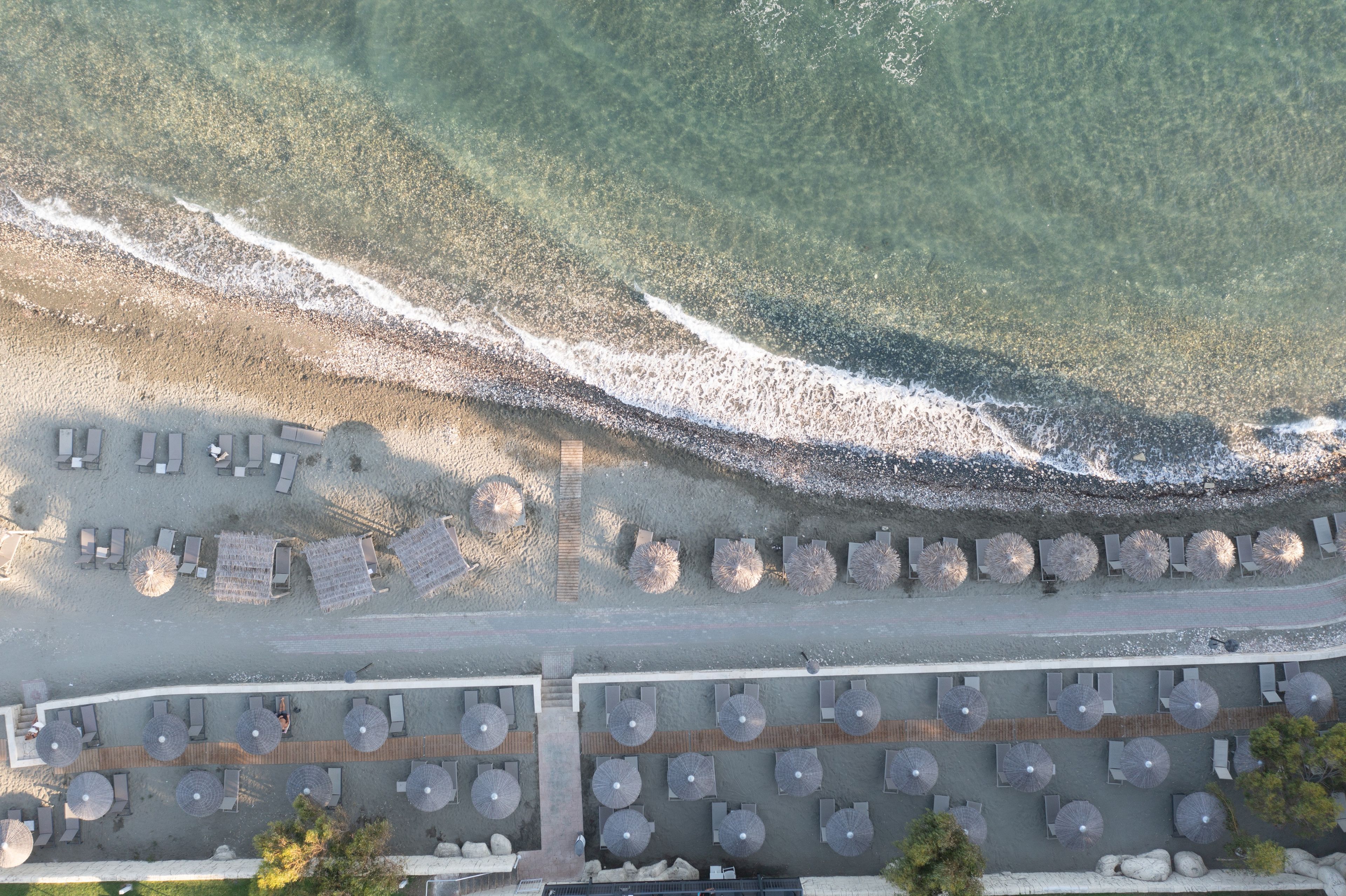An aerial view of a beachside hotel with rows of sun loungers and umbrellas, a wooden walkway, and a clear view of the ocean waves.
