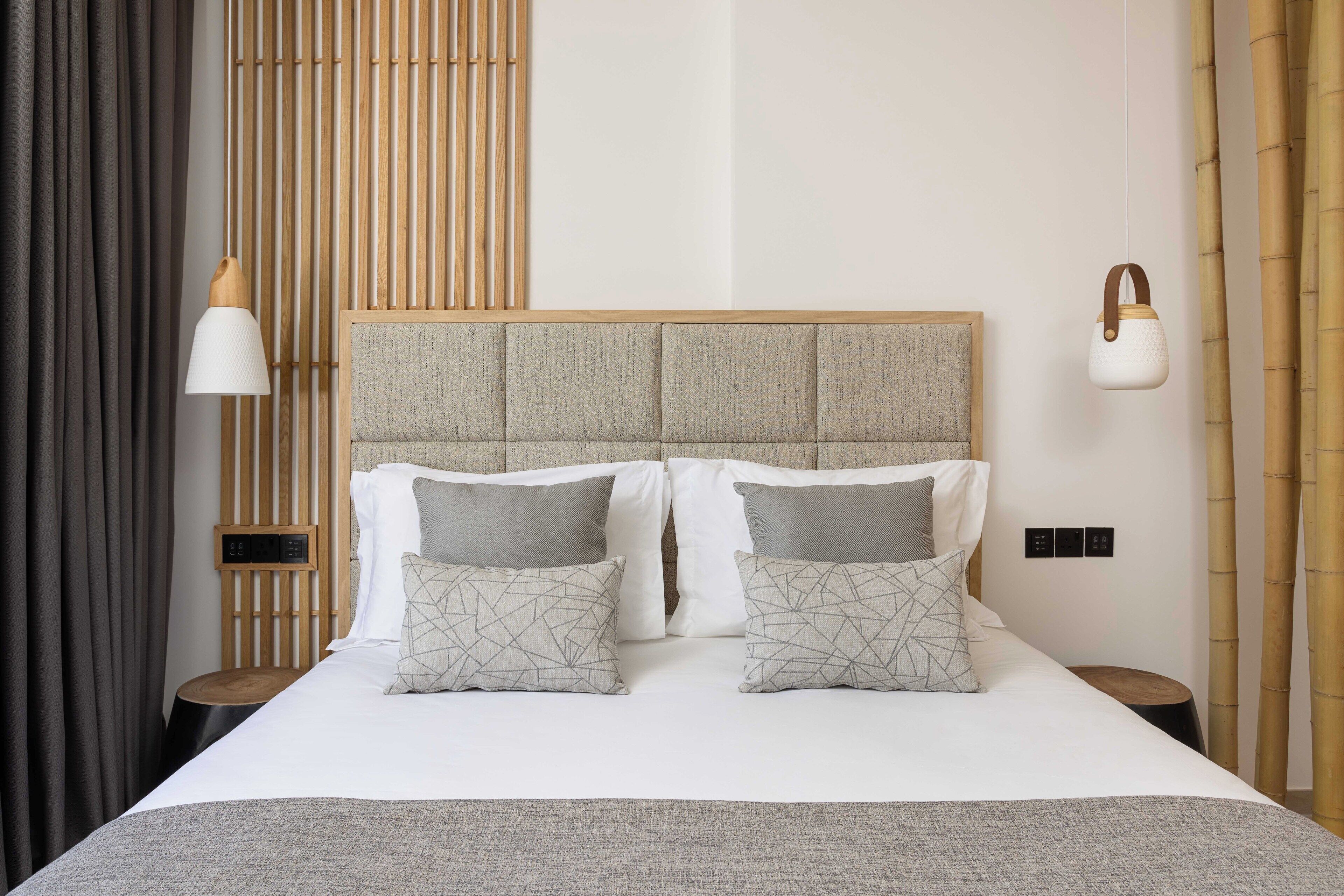 A neatly made bed with two pillows, a bedside table, a wall-mounted lamp, and a bamboo curtain.