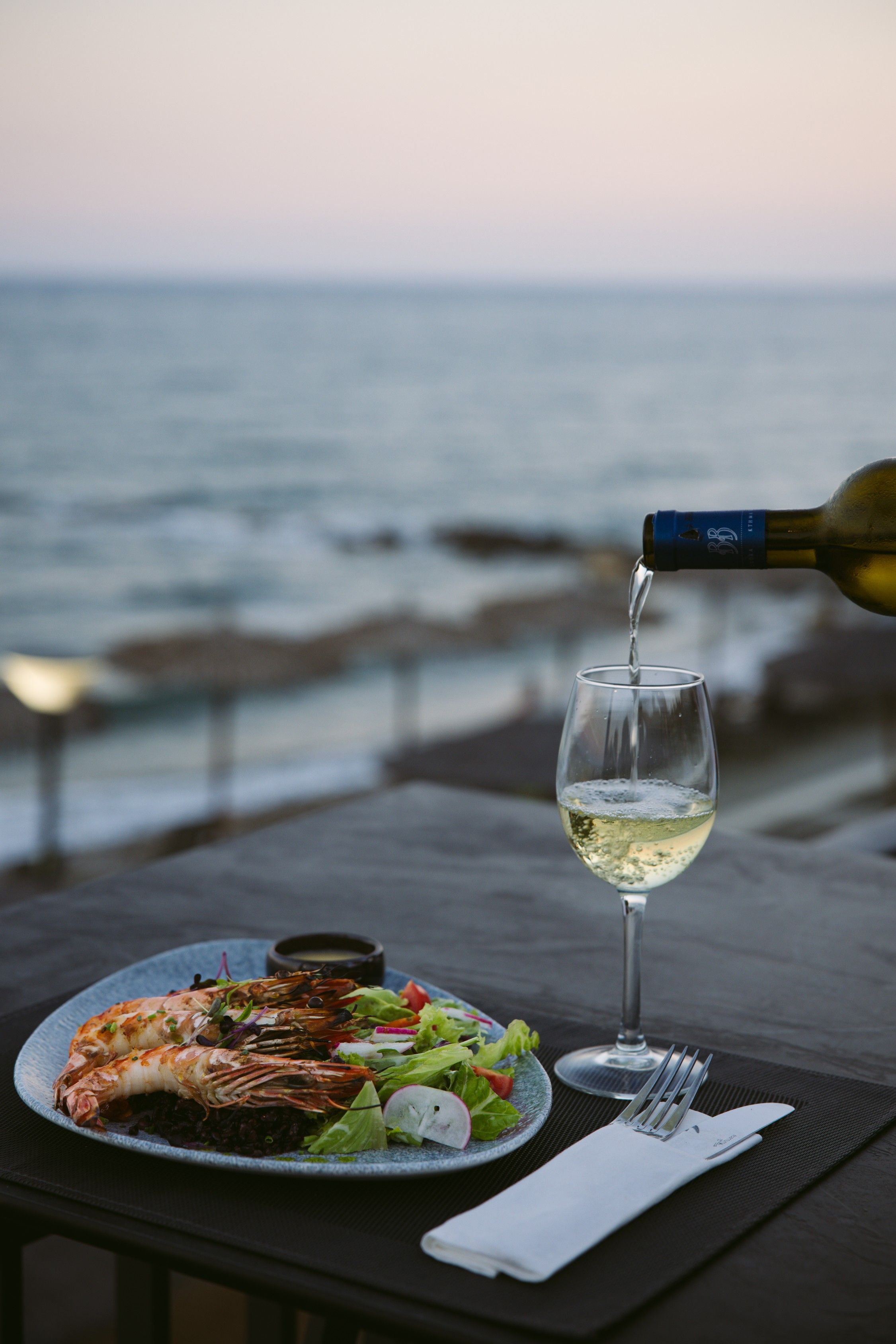 A table with a plate of food, a glass of white wine, and a bottle of wine being poured. The background features a view of the ocean.