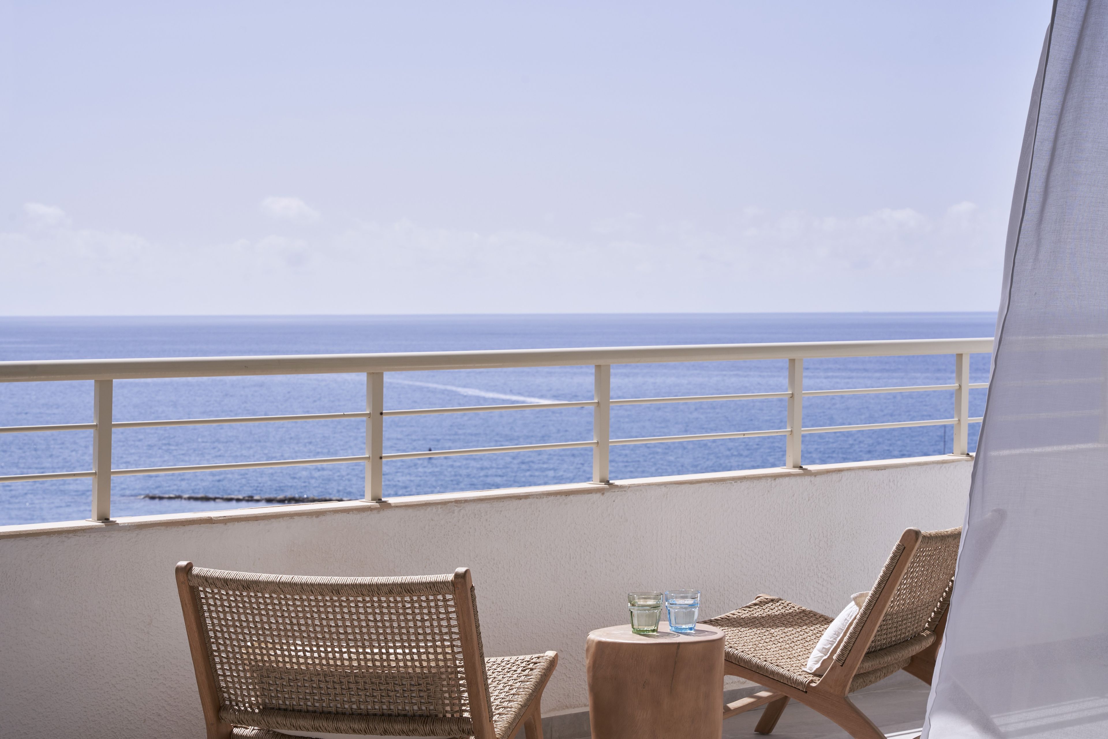 Un balcón con dos sillas de mimbre y una mesa redonda pequeña, con vista a un mar tranquilo.