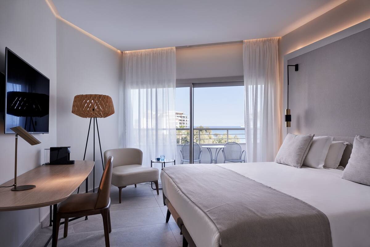 Een moderne hotelkamer met een groot bed, een bureau met lamp, een stoel en een televisie. Er is een balkon met een tafel en stoelen.