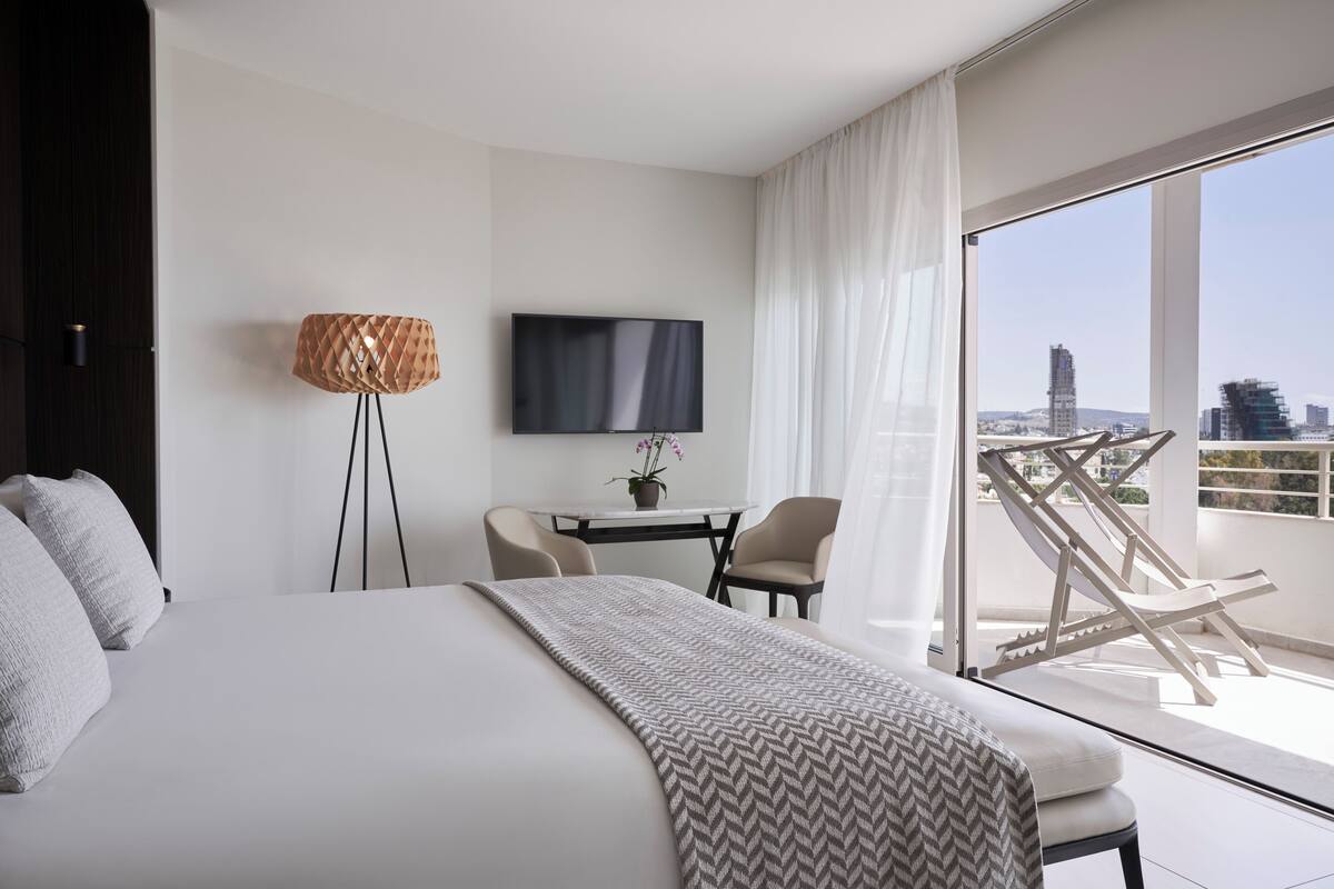 Een moderne hotelkamer met een groot bed, een aan de muur bevestigde televisie, een bureau met stoelen en een balkon met uitzicht op de stad.