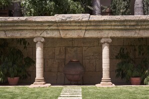 Una estructura de piedra con columnas y un gran jarro, rodeada de césped y plantas en macetas.