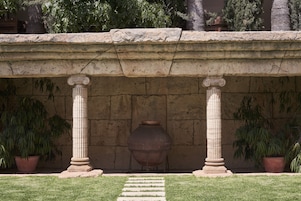 Una estructura de piedra con columnas y un gran jarro, rodeada de césped y plantas en macetas.