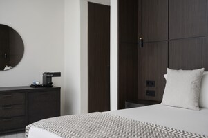 Habitación de hotel moderna con cama, mesita de noche, cafetera, espejo redondo y pared de madera oscura.