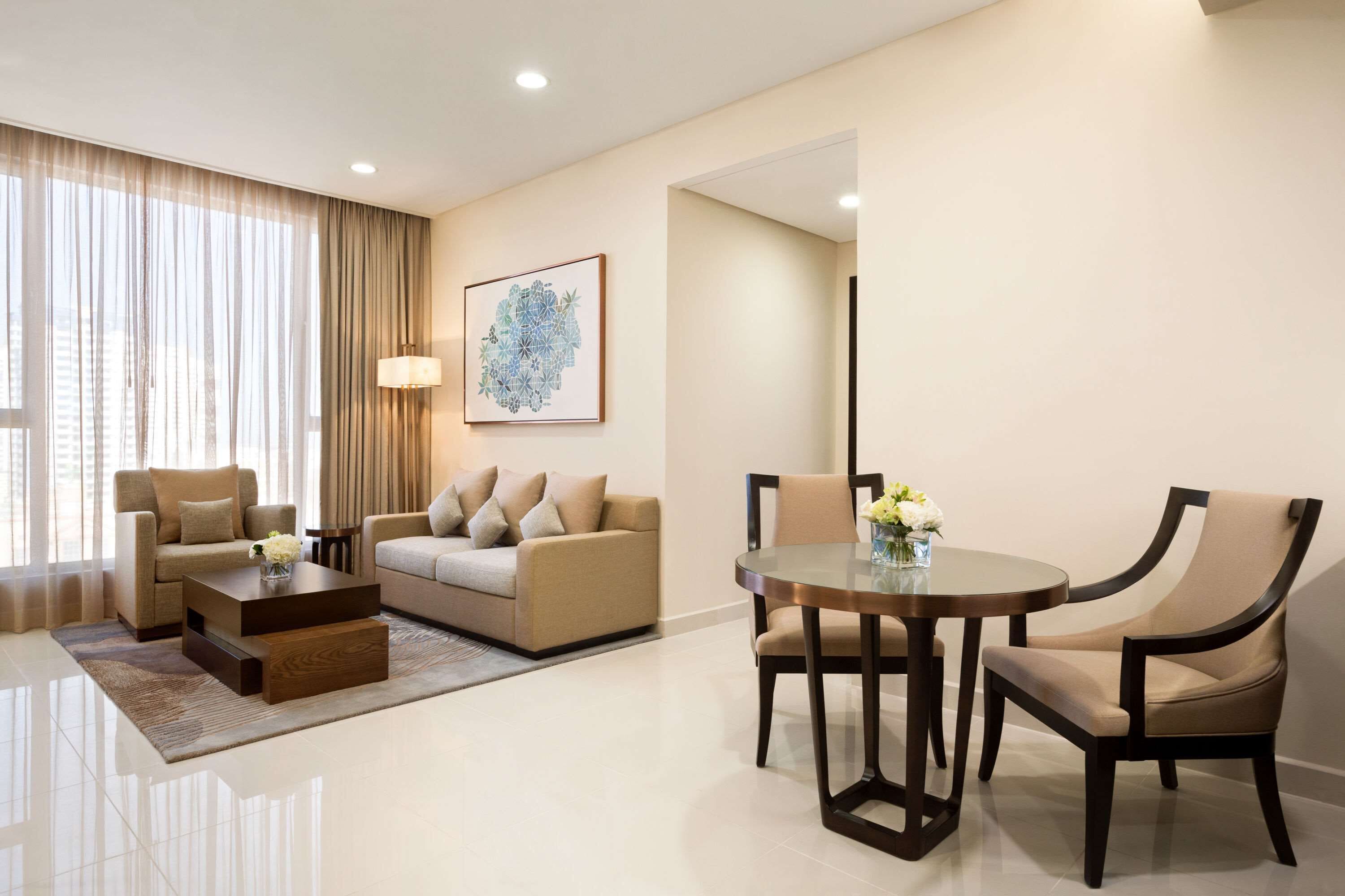 A modern hotel room with a sofa, a dining table with chairs, a coffee table, and a large window with curtains.
