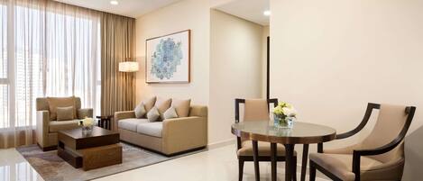 A modern hotel room with a sofa, a dining table with chairs, a coffee table, and a large window with curtains.