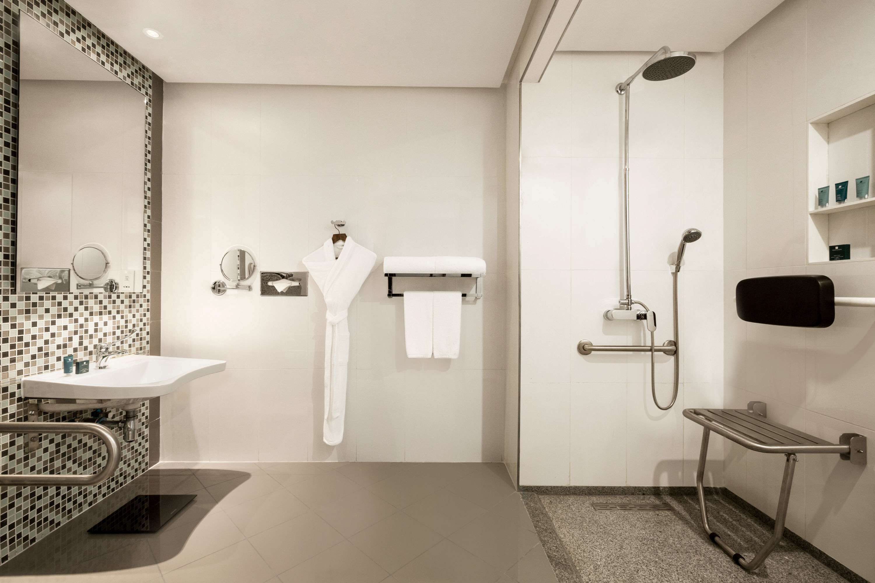 Un baño moderno con un lavamanos blanco, un espejo, un perchero para toallas, una ducha con cabezal de ducha de mano y una barra de apoyo.