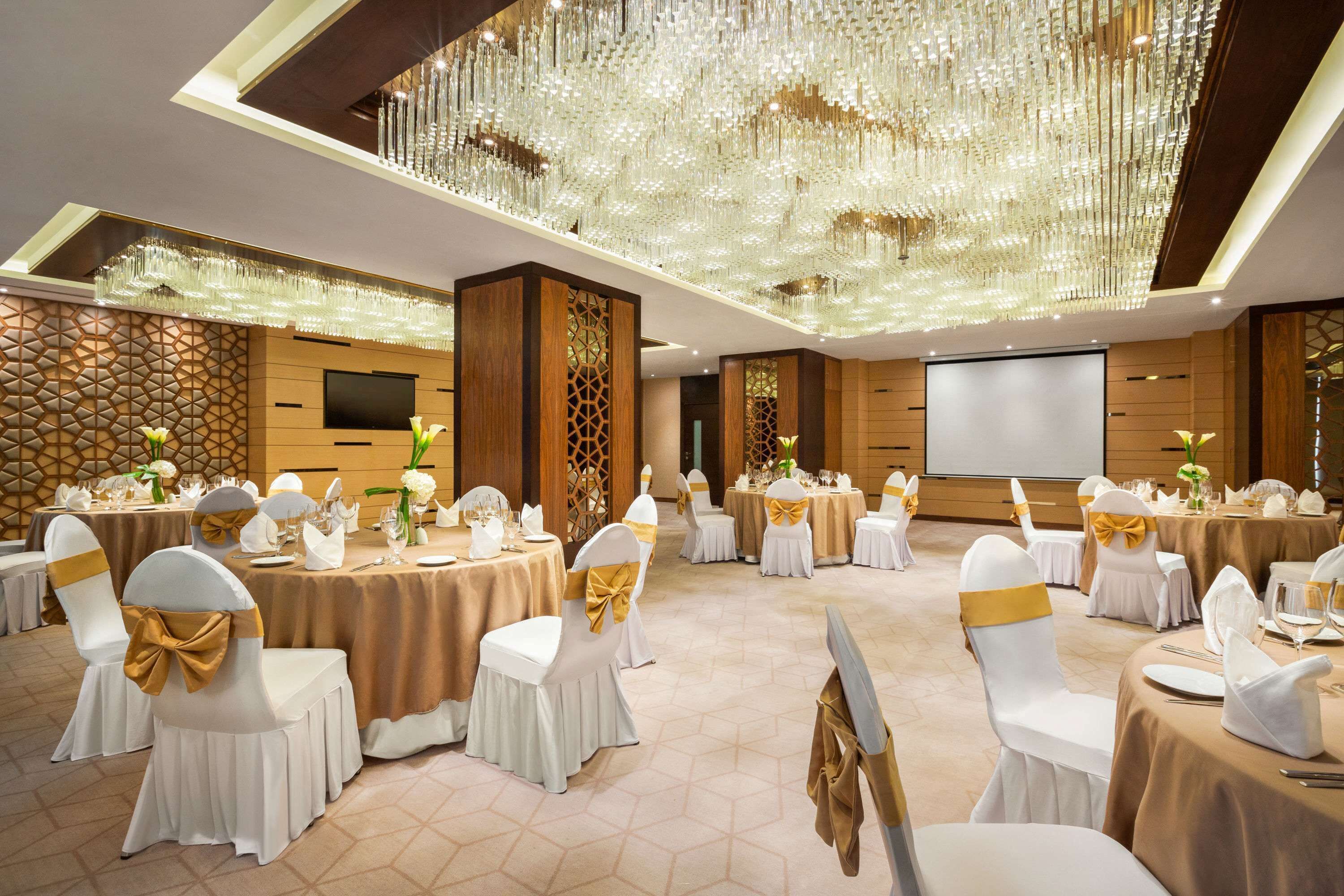 A banquet hall with round tables, white chairs with gold sashes, and a large chandelier.