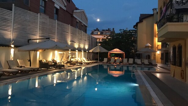 A pool area with lounge chairs and umbrellas, surrounded by buildings with balconies and a clear sky. - T & G Apartments (Marmaris)