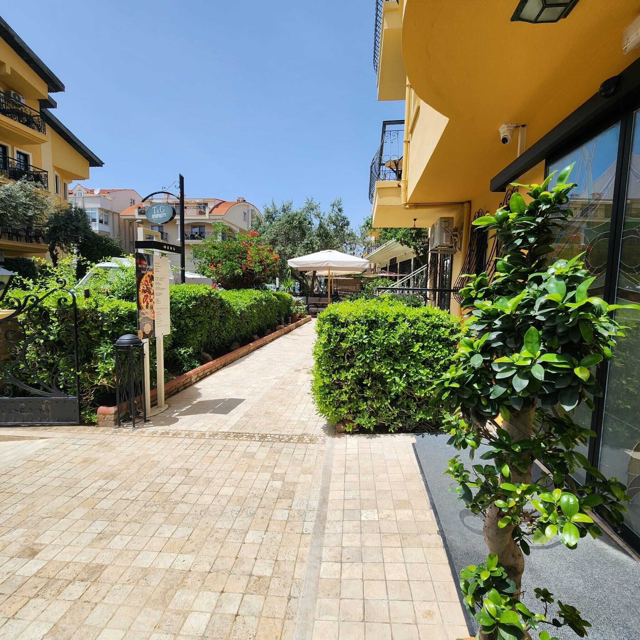 A paved pathway leading to a building with a signboard, surrounded by greenery and a clear sky.