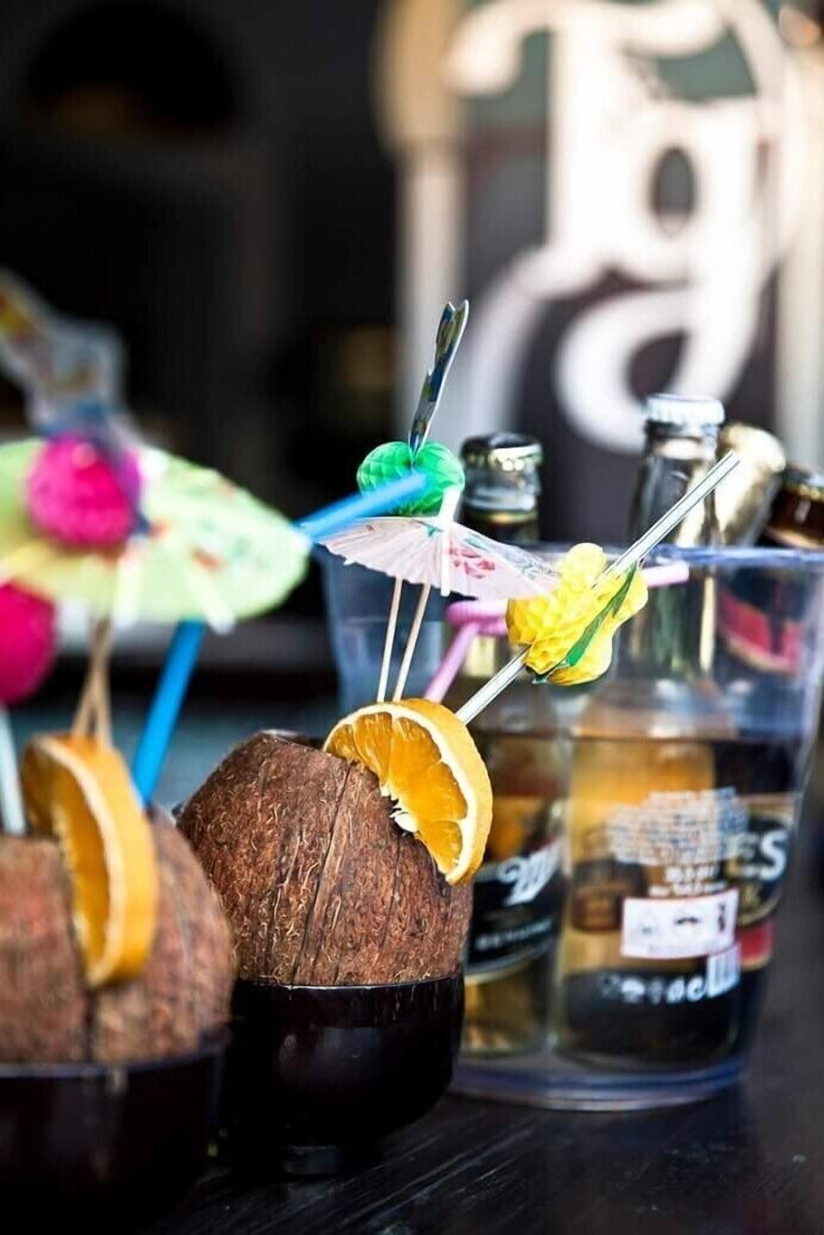 A bar area with coconut drinks garnished with fruit slices and colorful umbrellas, bottles of liquor, and a blurred background.