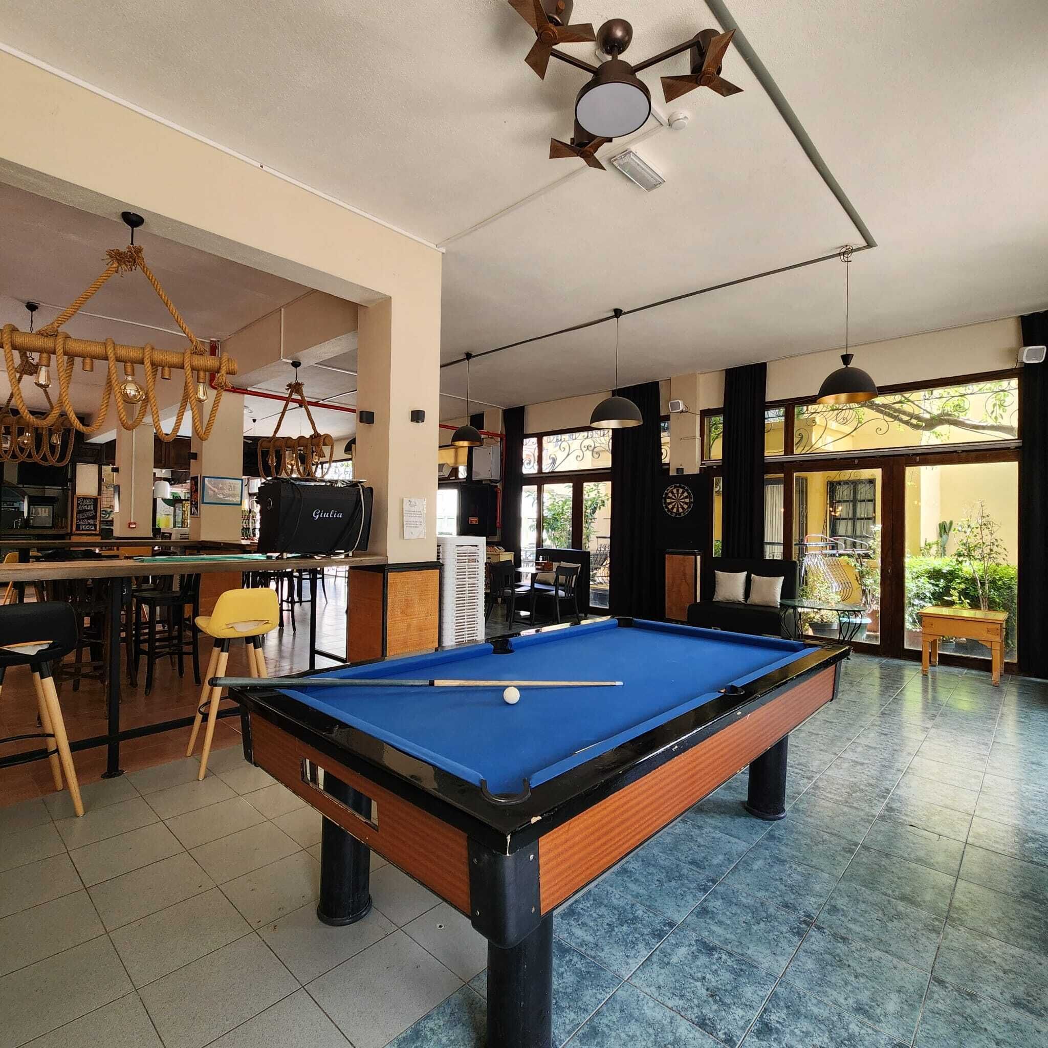 Une table de billard dans une pièce qui comprend un bar, un ventilateur de plafond et de grandes fenêtres offrant une vue sur l’extérieur.