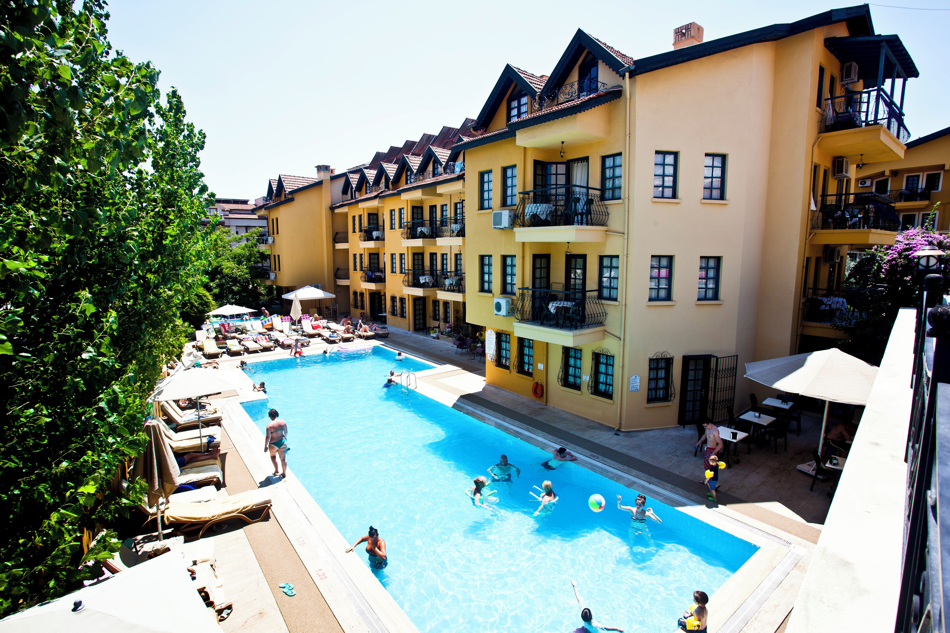 A swimming pool surrounded by multi-story buildings with balconies, outdoor seating, and umbrellas.